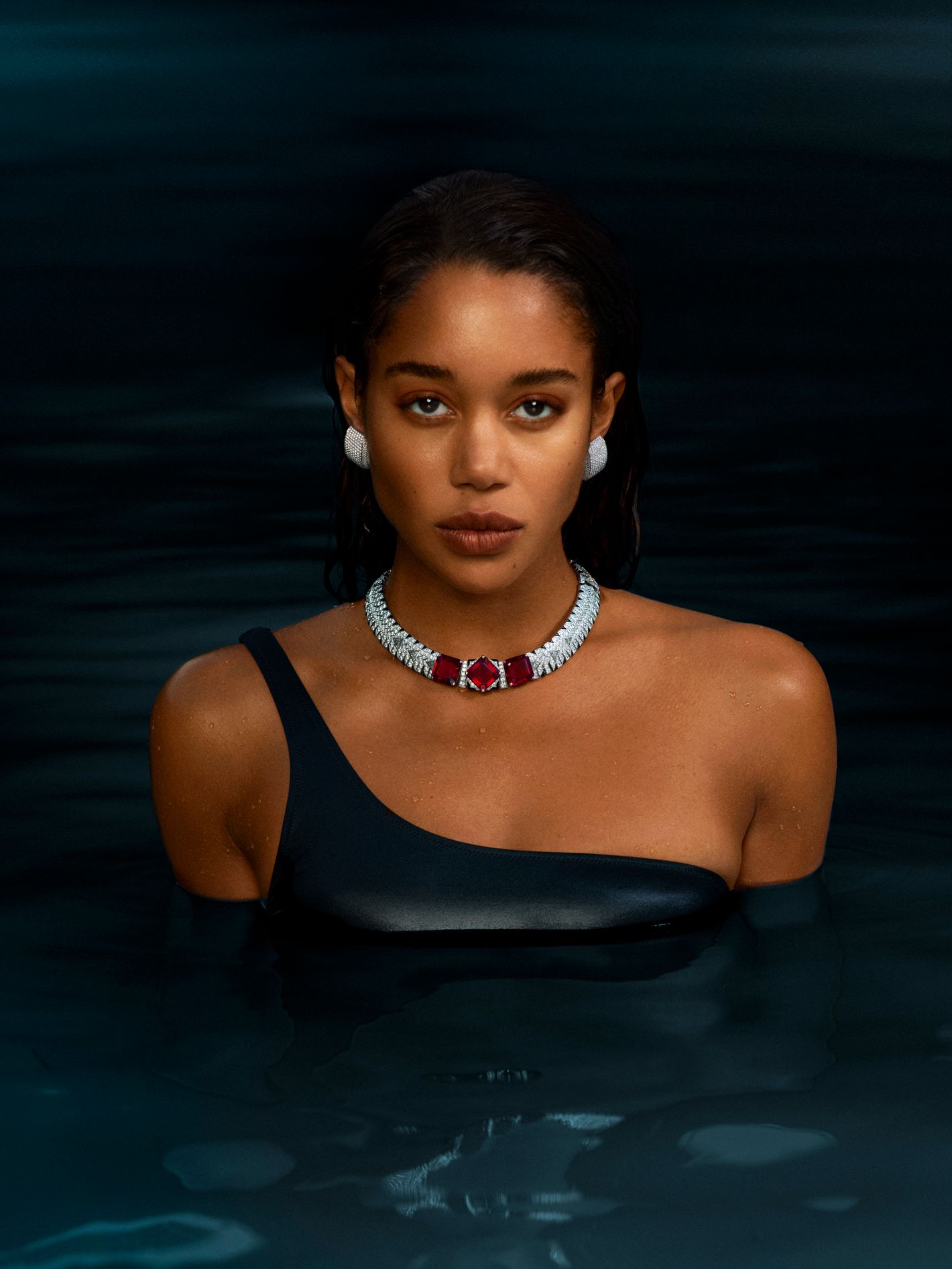 Adult female with dark hair and one shoulder swimsuit posing in water wearing a silver necklace with red gems and white stud earrings.