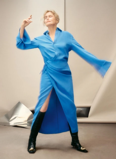 A mature woman in a blue wrap dress and black boots strikes a pose against a studio backdrop with foil accents, showcasing fashion.