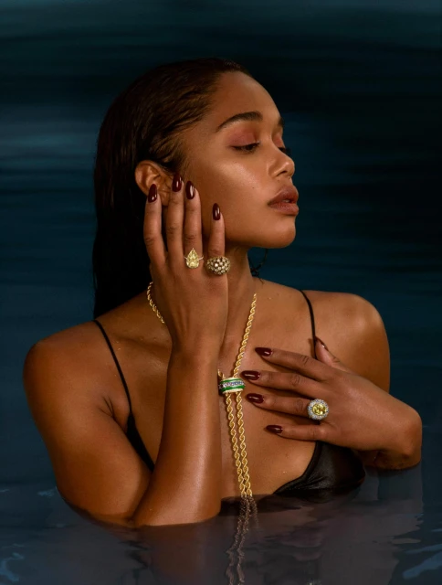 Woman in water wearing gold jewelry, including rings and a necklace, against a dark teal background with water reflections.