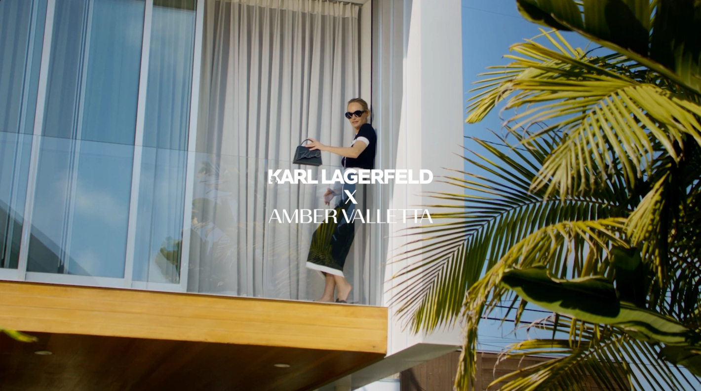 A woman poses on a balcony in a fashion shoot, holding a black purse, with palm trees and bright sky in the background and Karl Lagerfeld logo.