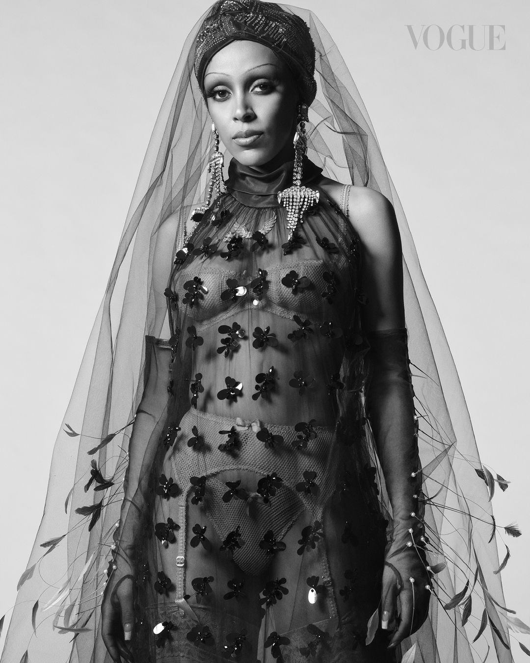 Black and white portrait of a stylish adult woman wearing a sheer floral dress, head wrap, veil, and statement earrings in a photo studio.