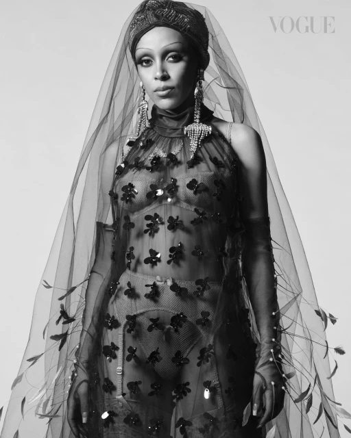 Black and white portrait of a stylish adult woman wearing a sheer floral dress, head wrap, veil, and statement earrings in a photo studio.