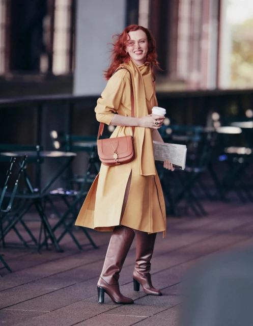 Smiling red-haired woman walks in a mustard-colored coat and brown boots, holding coffee and newspaper in an outdoor setting.