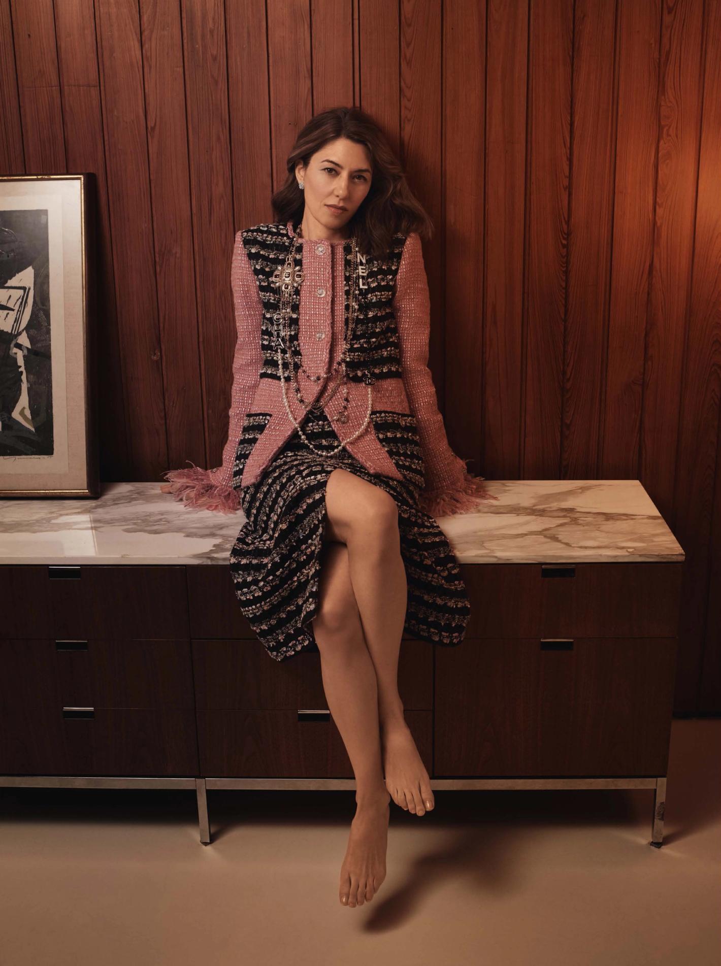 Adult female fashion model poses sitting on a wooden console in a studio wearing a pink-and-black tweed jacket and skirt.