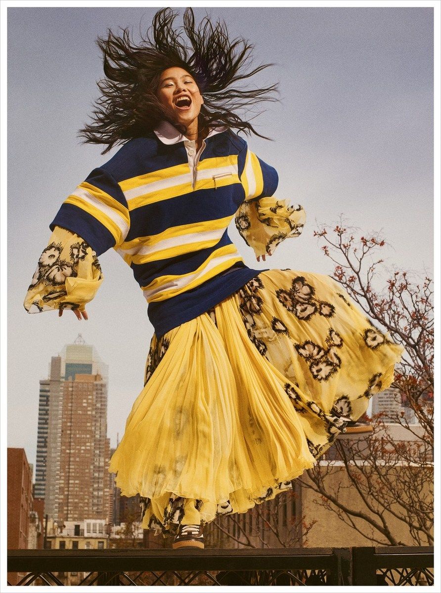 Model jumping in the air wearing a blue and yellow striped polo shirt and a pleated yellow floral skirt against a city sky background
