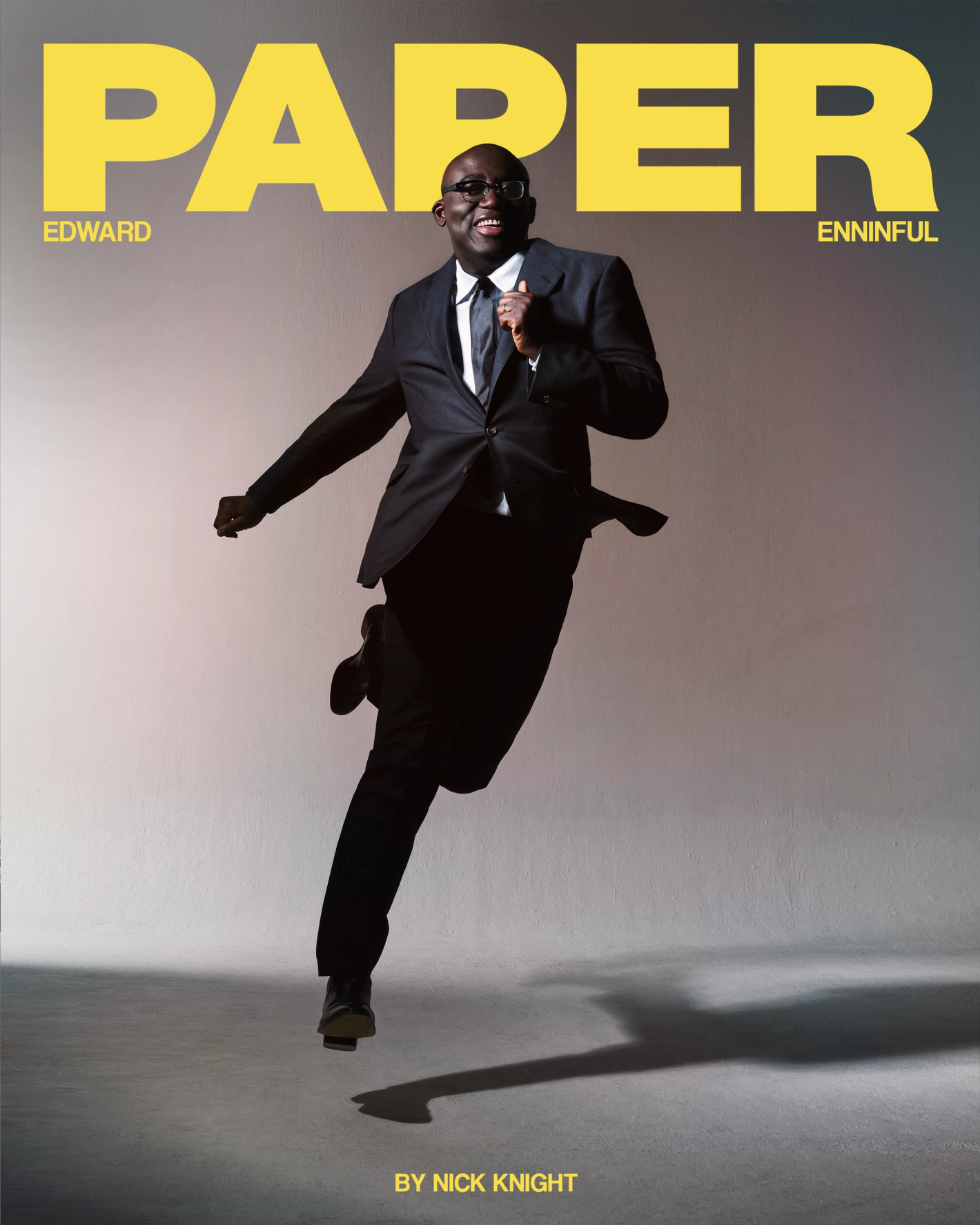 Edward Enninful leaping mid-air wearing a dark suit and tie against a plain grey studio backdrop with yellow Paper magazine masthead.