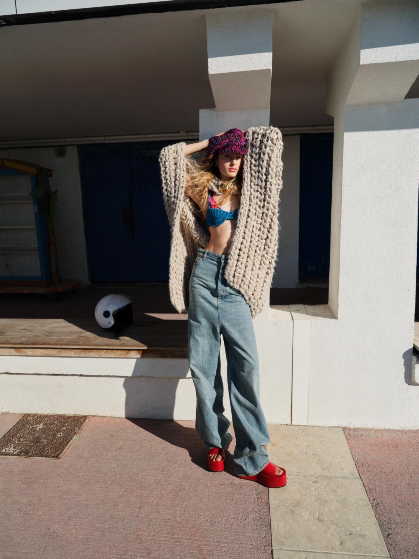 Fashionable young woman with long blonde hair poses on a sunlit porch in a knit outfit, hat, bikini top, red sandals, and yin yang helmet.