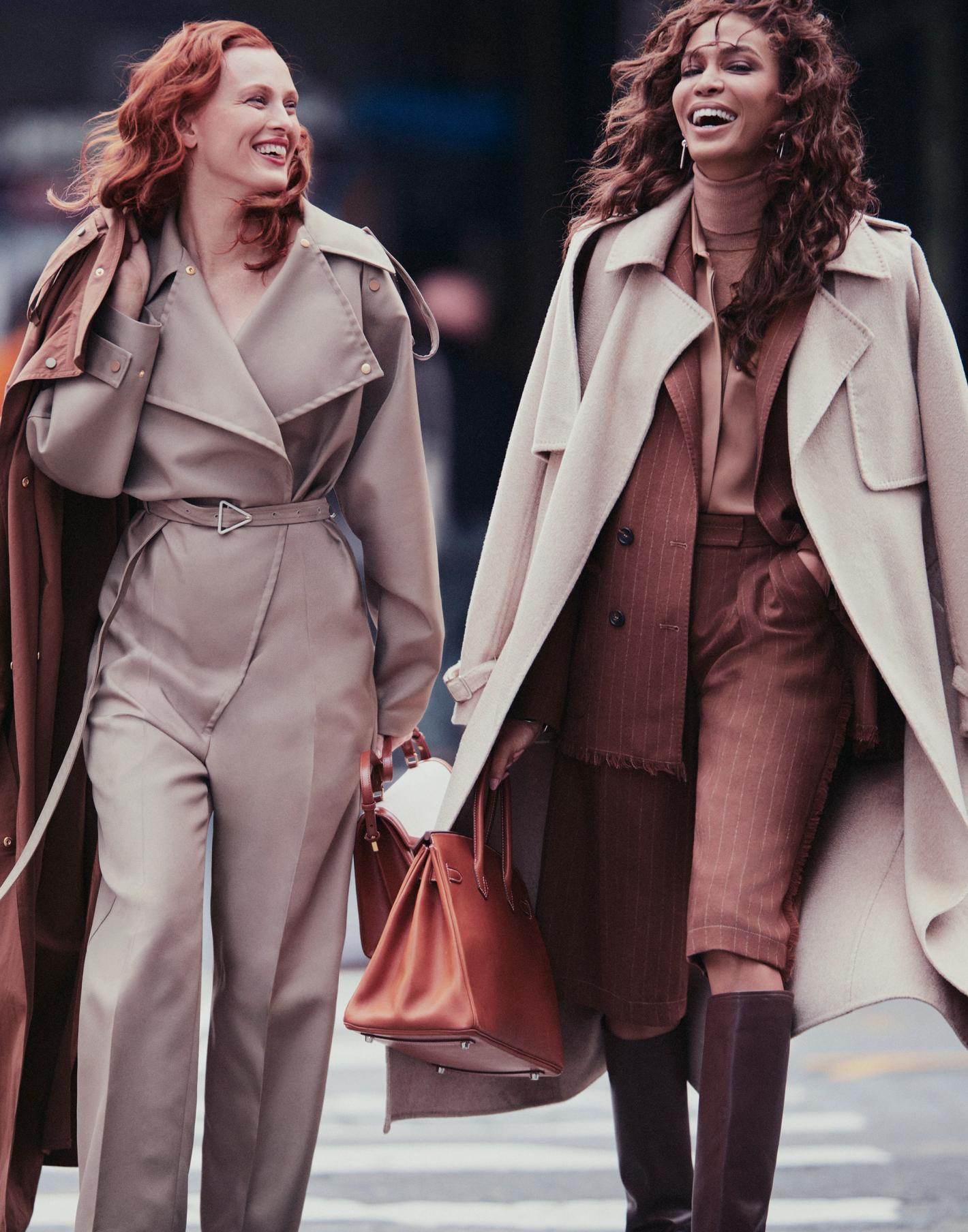 Two women in trench coats laugh while walking in a city. The woman on the right carries a brown leather handbag, and the sky is overcast.