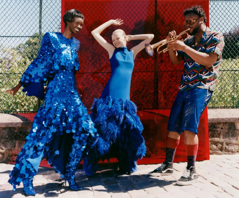 Three people in an outdoor setting: two adult women in blue gowns and a man playing a trumpet, standing in front of a colored backdrop.