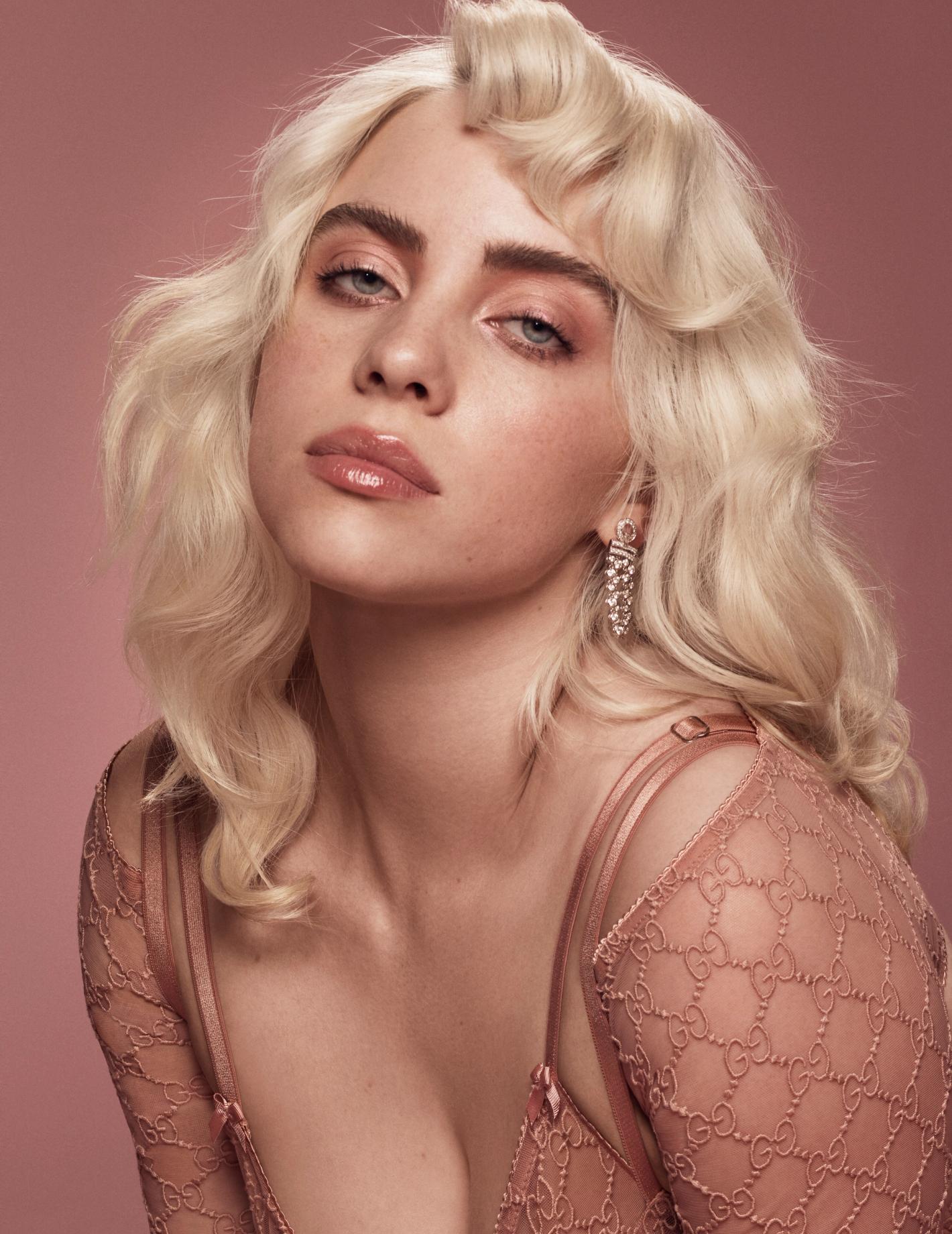 Close-up portrait of a young adult woman with platinum blonde wavy hair, pink lipstick, and pink Gucci-patterned top against a pink backdrop.