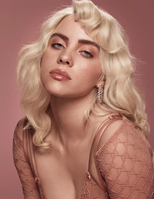 Close-up portrait of a young adult woman with platinum blonde wavy hair, pink lipstick, and pink Gucci-patterned top against a pink backdrop.