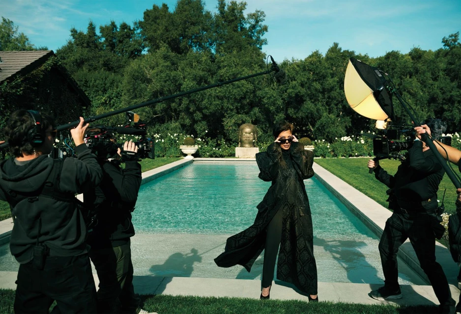 Fashion shoot outdoors featuring a model in a black coat, surrounded by camera crew near a swimming pool and green foliage.