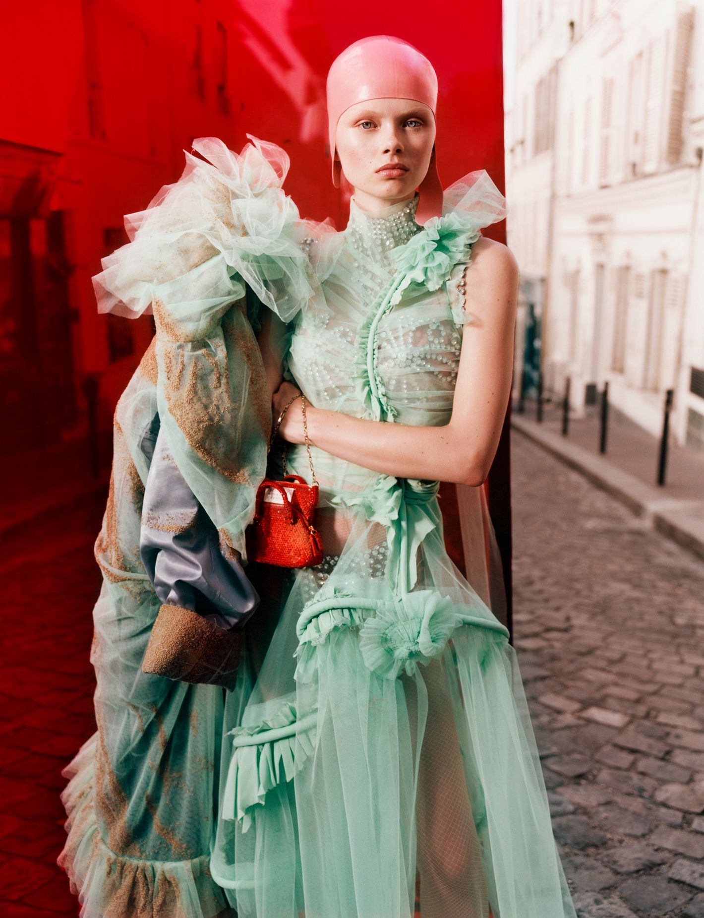 Fashion editorial shot featuring a female model in an avant-garde green tulle gown and pink cap, against a split red and street scene backdrop.