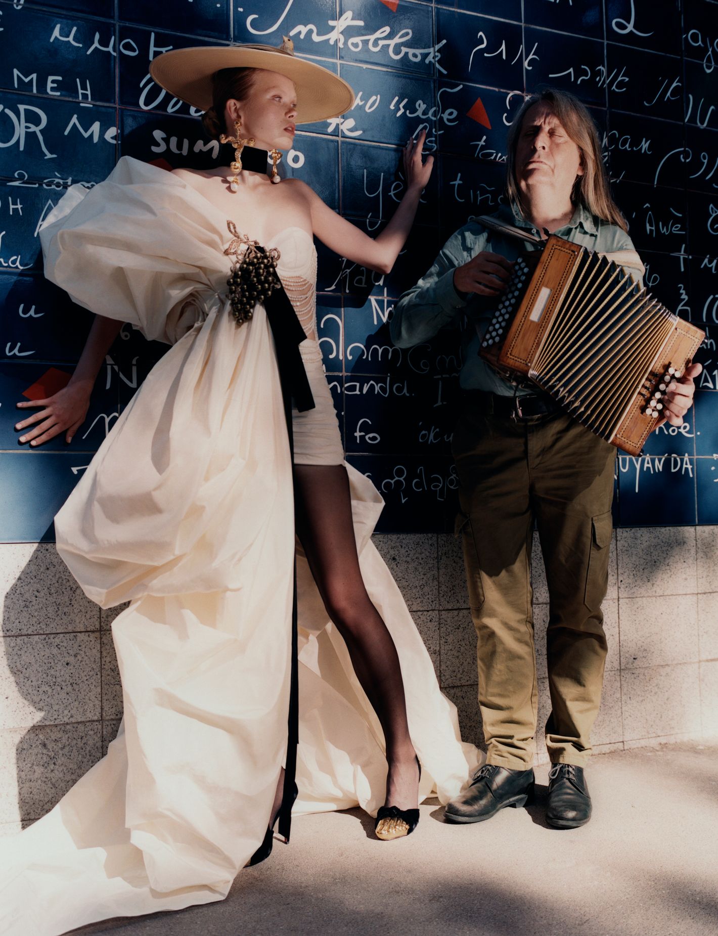 Elegant fashion model in haute couture gown poses with accordion player in an indoor location with dark blue textured walls.