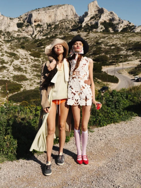 Two women pose outdoors near mountains. One wears a light dress and red shoes; the other, a coat, shorts, and hat in a fashion shot.