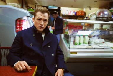 Taron Egerton wearing a navy pea coat seated inside a convenience store or deli with shelves and display cases behind him.