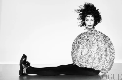 Model with voluminous dark curly hair wearing a spherical metallic top and black trousers seated on studio floor