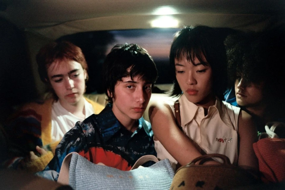 Group of four young adults sitting in a car, looking toward the camera with neutral expressions, illuminated by soft, natural light.