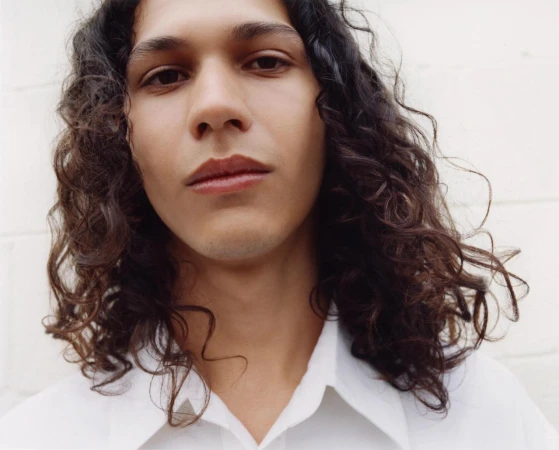 Close up portrait of a young man with long curly dark hair wearing a white shirt against a plain light wall.