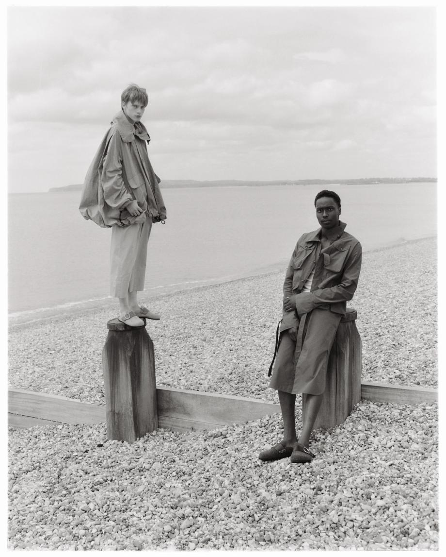Two fashion models pose in jackets and pants on a pebbled beach with a sea background, showcasing a neutral color palette and outdoor setting.