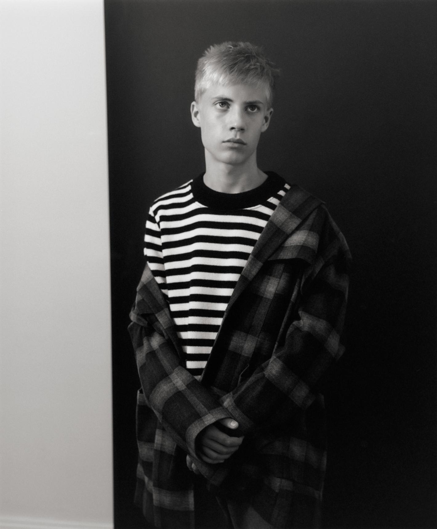 Young man with short blonde hair wearing a striped crewneck sweater and plaid jacket against a split backdrop