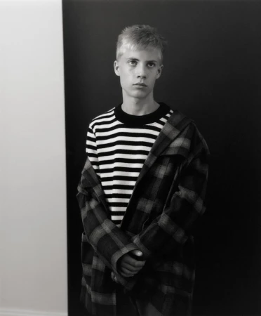 Young man with short blonde hair wearing a striped crewneck sweater and plaid jacket against a split backdrop