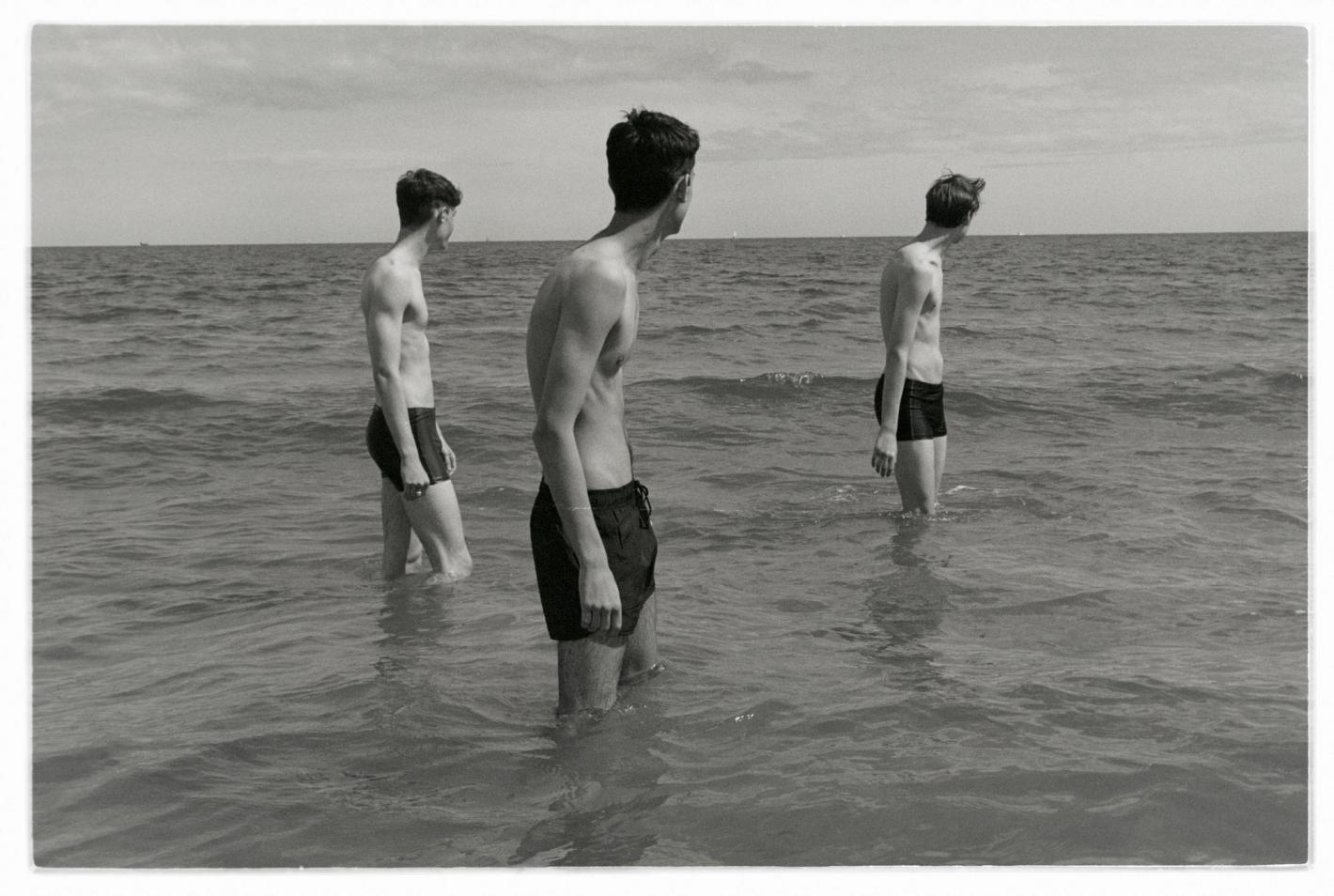 Three shirtless young men stand in ocean surf on a sunny day, facing away from the camera in a black and white photograph.