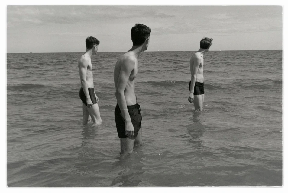 Three shirtless young men stand in ocean surf on a sunny day, facing away from the camera in a black and white photograph.