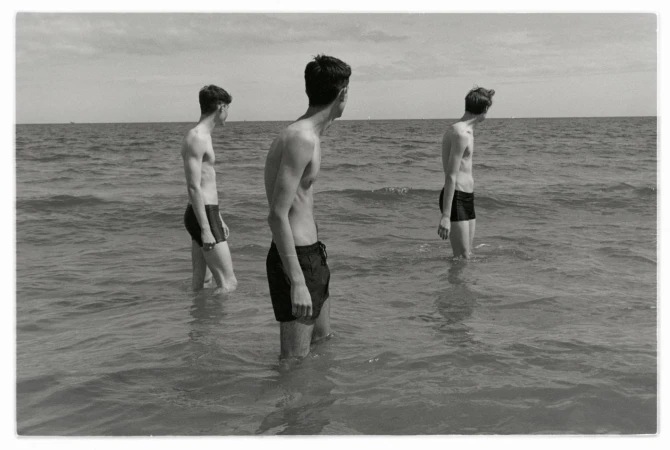 Three shirtless young men stand in ocean surf on a sunny day, facing away from the camera in a black and white photograph.