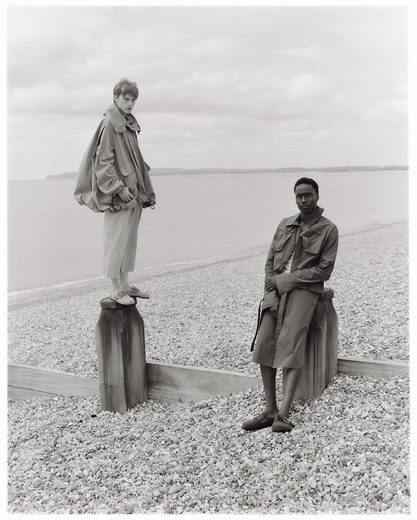Two fashion models pose on a pebble beach near the ocean on an overcast day, showcasing neutral-toned coats and relaxed poses in a monochromatic scene.