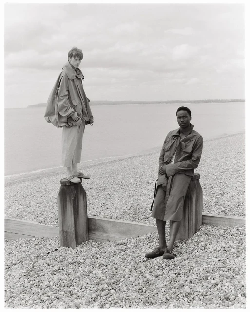 Two fashion models pose on a pebble beach near the ocean on an overcast day, showcasing neutral-toned coats and relaxed poses in a monochromatic scene.