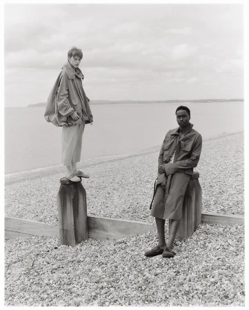 Two fashion models pose on a pebble beach near the ocean on an overcast day, showcasing neutral-toned coats and relaxed poses in a monochromatic scene.