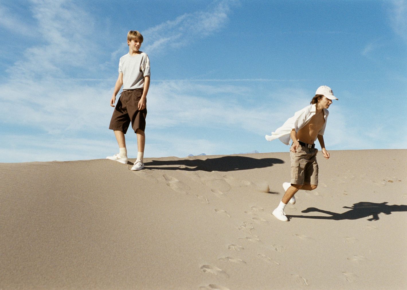 Two kids on a sand dune under a blue, cloudy sky. One stands on the dune's crest, and the other runs down, creating a dynamic visual.