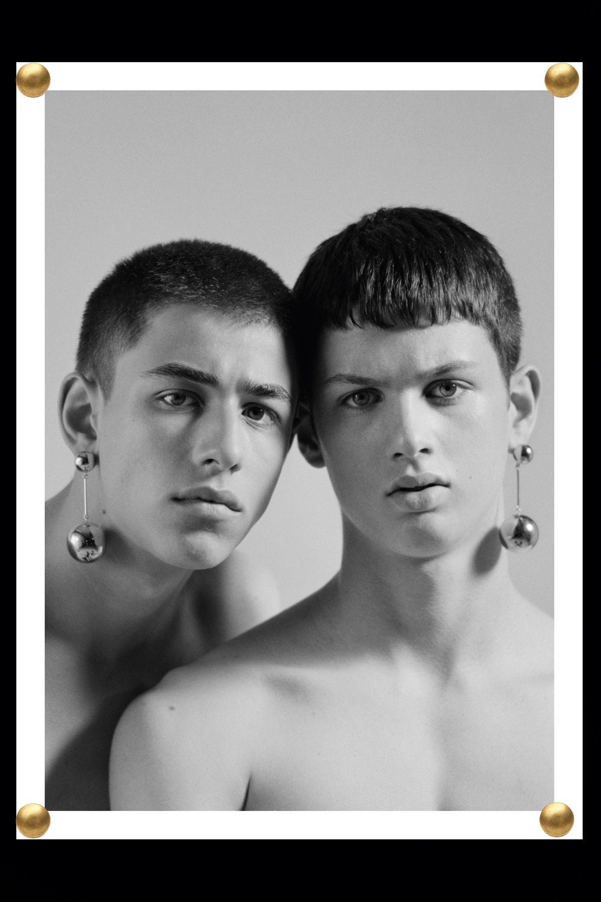 Black and white close-up of two adult male models with bare chests and short haircuts, wearing earrings, against a studio backdrop.
