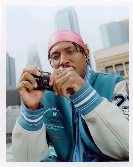 Man with pink headscarf holds a camera in an urban setting with tall buildings and overcast lighting, wearing an Amiri varsity jacket.