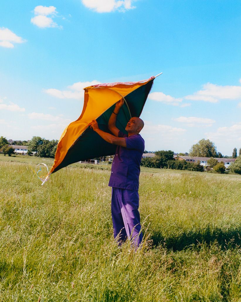 Adult male in purple leisure suit standing in a field of grass, holding a yellow, orange, and black diamond-shaped kite on a bright day.
