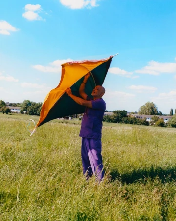 Adult male in purple leisure suit standing in a field of grass, holding a yellow, orange, and black diamond-shaped kite on a bright day.
