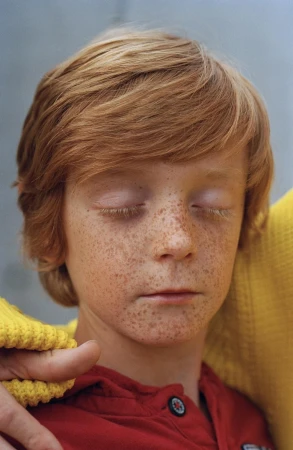 Close-up portrait of a young person with red hair, closed eyes, and visible freckles wearing a red shirt and yellow knitted cardigan