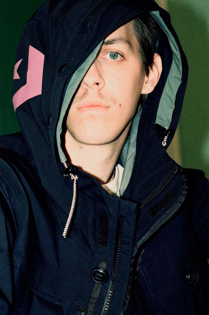 Close-up shot of a fair-skinned adult male with light eyes and a mustache wearing a hooded blue jacket with a pink graphic.