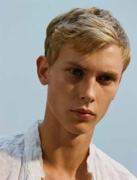 Portrait of a young adult male with blond hair wearing a white linen shirt, set against a soft blue background in natural light.