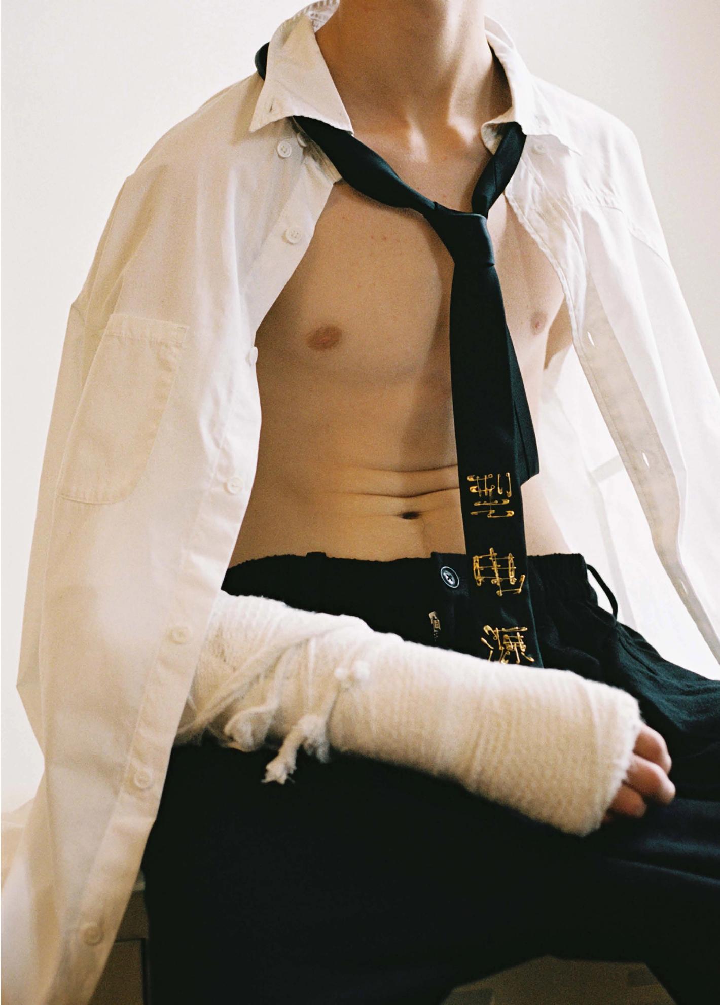 Man with an arm cast, wearing a white shirt and tie, sits in a studio with a neutral background, showcasing a blend of fashion and healthcare themes.