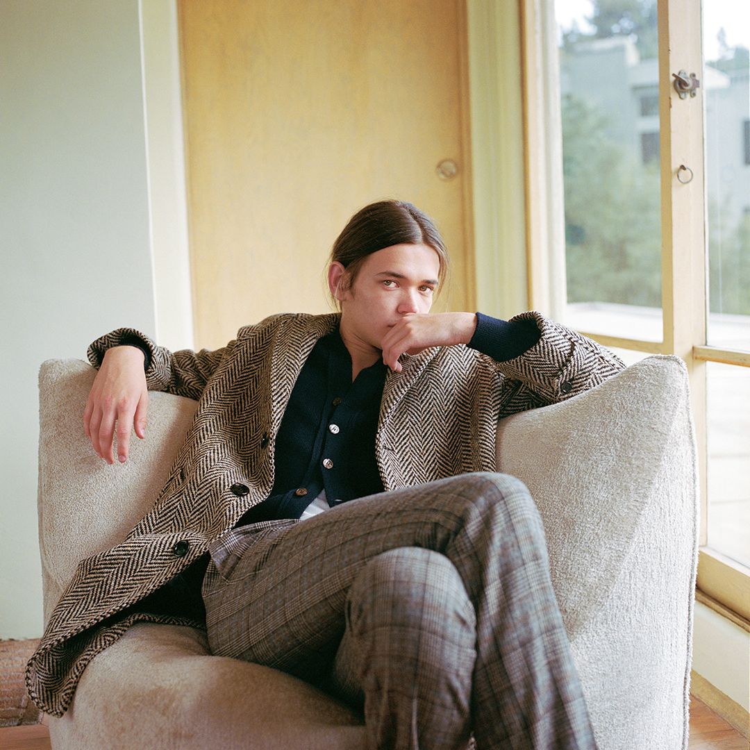 A young adult male with long brown hair is posed sitting in an armchair, wearing a herringbone overcoat and plaid trousers in a relaxed indoor setting.