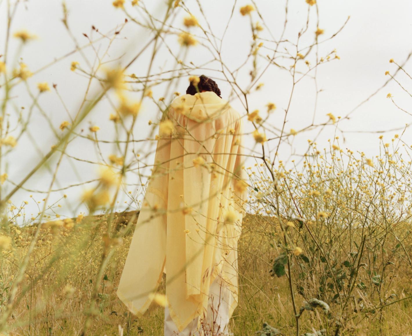 Woman in yellow hooded cape standing in a field of small yellow wildflowers, back to camera, rural outdoor setting with overcast sky.