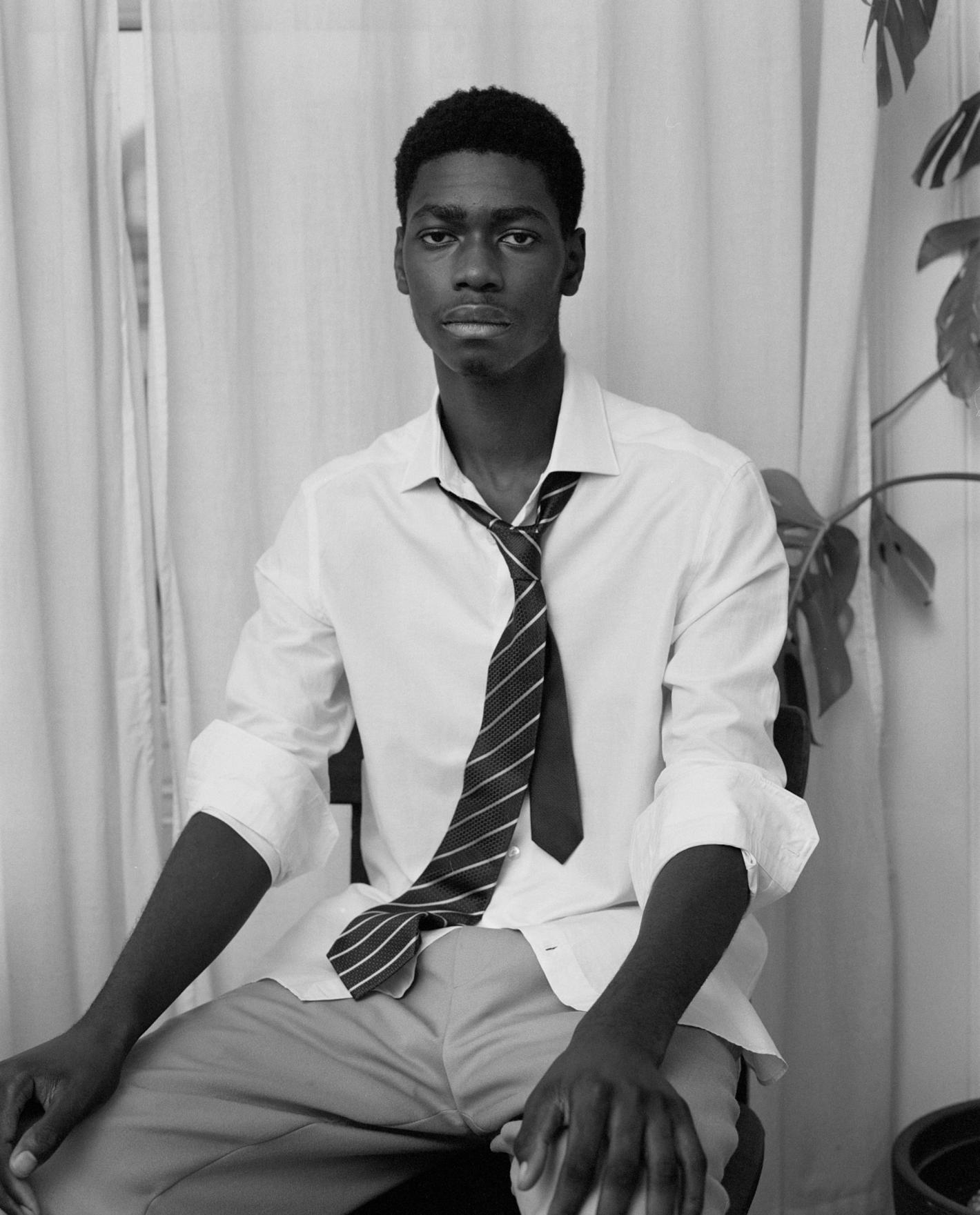 Portrait of an African-American man in a white shirt and tie seated indoors with a serious expression in front of a curtain and houseplant.