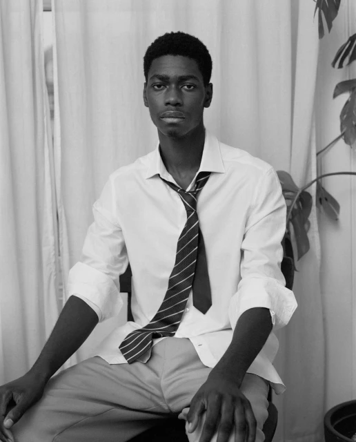 Portrait of an African-American man in a white shirt and tie seated indoors with a serious expression in front of a curtain and houseplant.