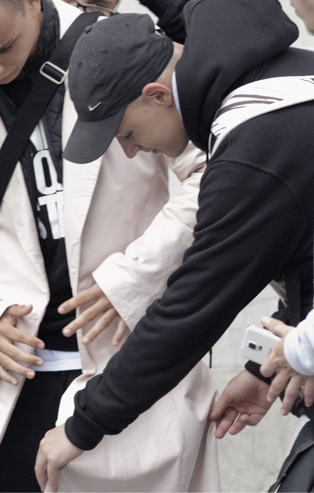 A group of people surround an adult male outdoors, dressed in a light coat, black hoodie, and baseball cap with a visible logo.
