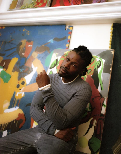 Adult male with braided hair sits in front of large, colorful abstract paintings in an indoor environment, wearing a gray sweater.