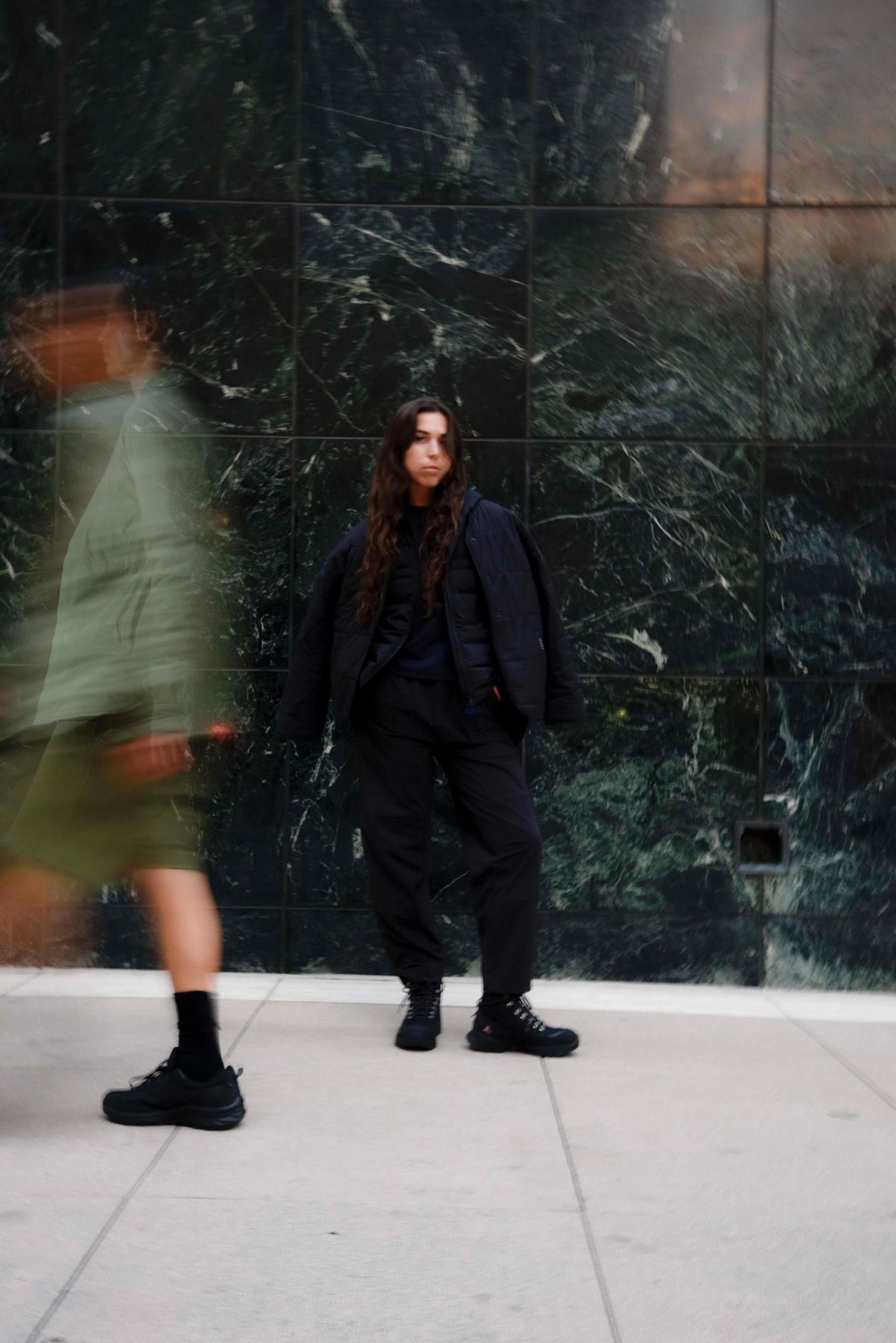 Woman in black outfit poses in front of a marble wall with a blurred figure walking by in an outdoor urban setting.