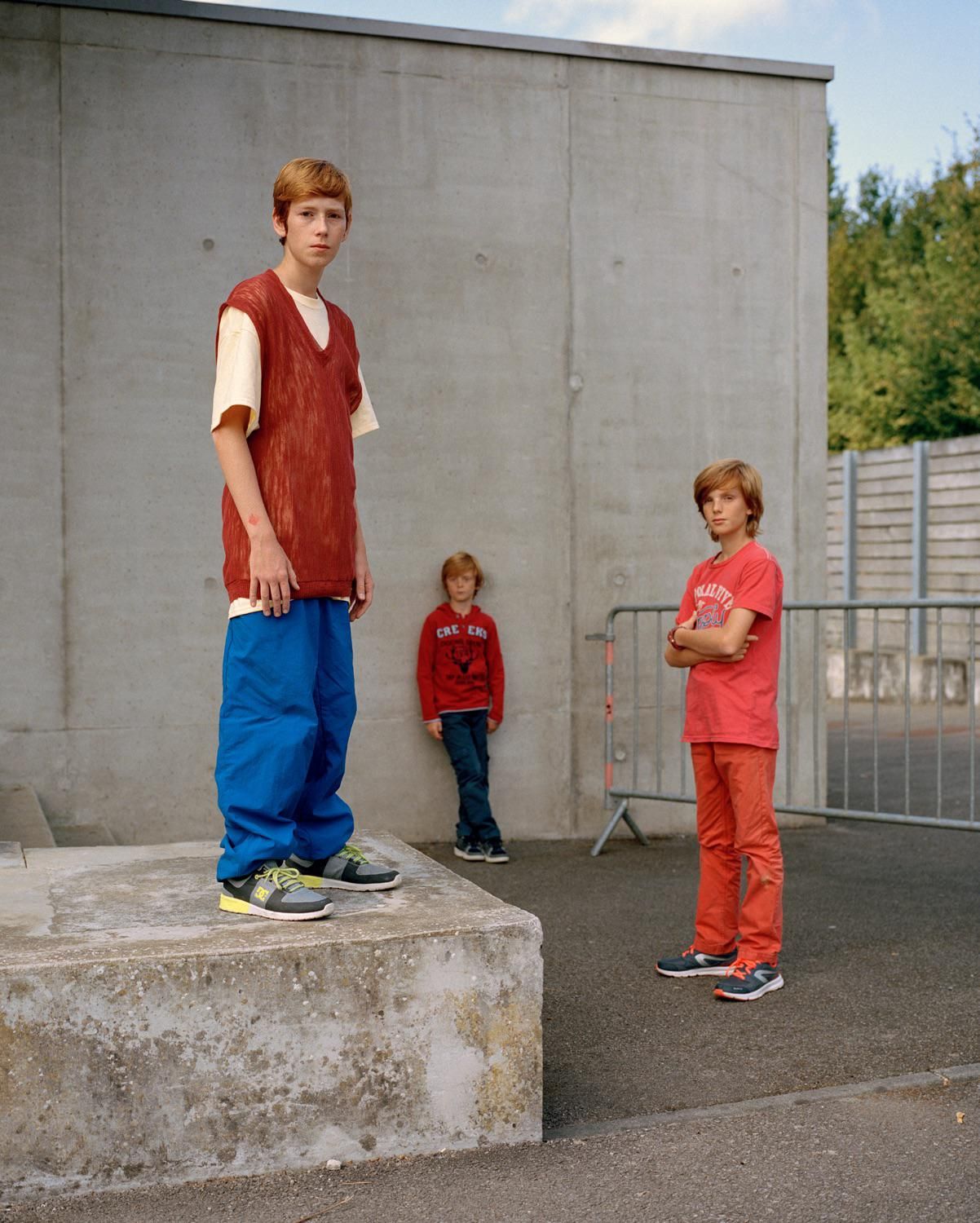 Three red-haired children in casual clothing stand outdoors against a grey concrete wall. One child stands on a concrete block.