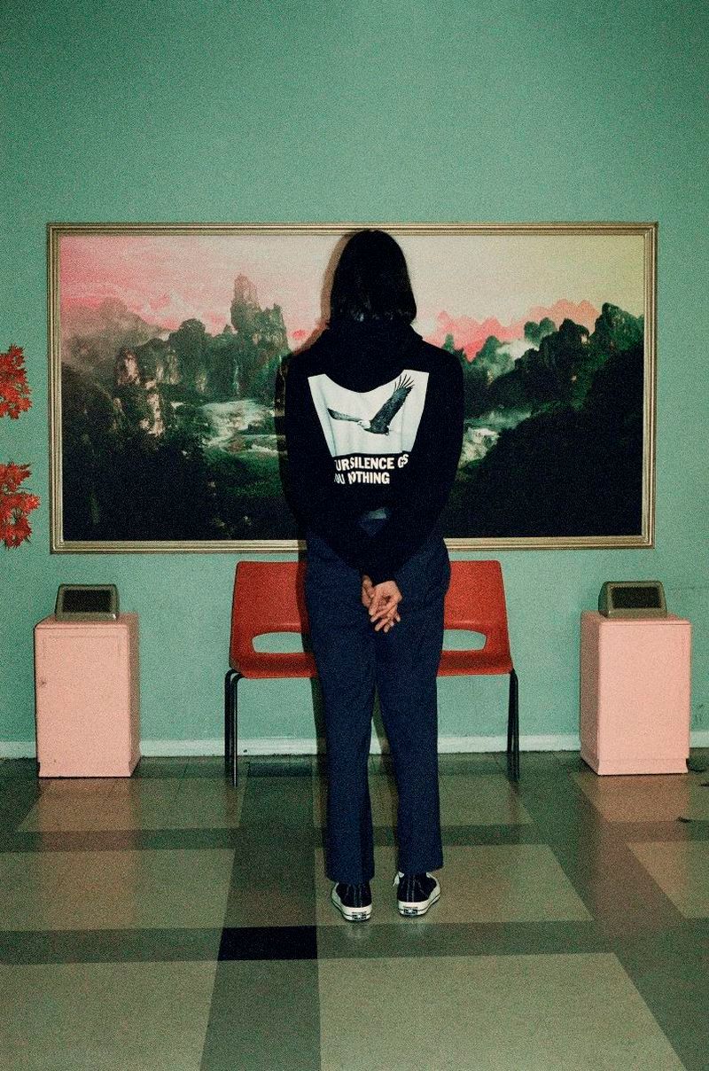Back view of a person in a hoodie standing in front of a landscape painting in a room with tiled floors, pale green walls, and furniture.